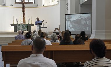 Das Foto zeigt mehrere Personen auf Kirchenbänken von hinten, während sie einem Vortrag zur Ausstellung 'Auf der Flucht. Frauen und Migration' in der Johanneskirche zuhören © Landeshauptstadt Düsseldorf