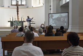 Das Foto zeigt mehrere Personen auf Kirchenbänken von hinten, während sie einem Vortrag zur Ausstellung 'Auf der Flucht. Frauen und Migration' in der Johanneskirche zuhören © Landeshauptstadt Düsseldorf