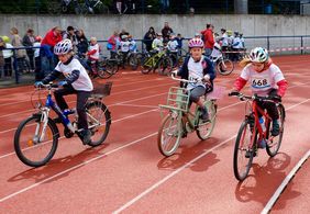 Foto von 3 Kindern auf Fahrrädern beim Fahrradrennen, im Hintergrund Zuschauer.