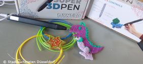 Foto mit 3D-Druck-Stift, einem gebastelten Dino, Filament in verschiedenen Farben und einem Buch mit Anleitungen