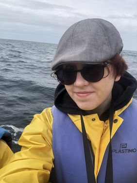Kim Zettl auf einem Expeditionsboot. Kim trägt Mütze und Sonnenbrille, eine gelbe Regenjacke, darüber eine Schwimmweste.