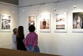 Das Foto zeigt drei Personen von hinten, während sie fünf Bilder der Ausstellung 'Auf der Flucht. Frauen und Migration' in der Johanneskirche betrachten © Landeshauptstadt Düsseldorf