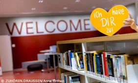 Foto der Zentralbibliothek Düsseldorf im KAP1 mit einem Bücherregal und der Wandschrift "Welcome". Jemand hält ein gelbes Herz aus Pappe mit der Aufschrift "Ich höre dir zu!" auf das Regal.