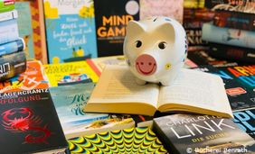 Fotografie mit vielen Büchern auf denen ein Sparsschwein aus Porzelan steht.