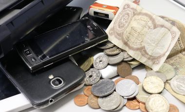 Mehrere Smartphones mit zerbrochenen Displays, Druckerpatronen sowie Münzen und Scheine aus verschiedenen Ländern auf einem Schreibtisch