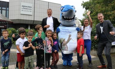 Josef Hinkel (Bürgermeister Düsseldorf), Silke Heise (Weltretterbande) und Dr. Jochen Reiter (Aquazoo-Direktor) inmitten einer Kindergruppe vor dem Gebäude des Aquazoo Löbbecke Museum. Ein Hai (Maskottchen) hält das Poster zum Aquazoo-Familienfest in den Flossen.